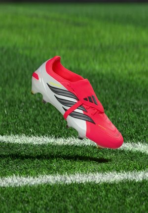 Roter und weißer Fußballschuh mit schwarzen Streifen schwebt über grünem Gras nahe einer weißen Spielfeldlinie.