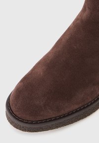 Close-up da ponta de uma bota de camurça castanha com sola de borracha texturizada castanha escura e costura visível em fundo branco.