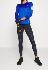 Sininen pitkähihainen suorituspaita, jossa on valkoiset Adidas-raita, yhdistetty mustiin legginsseihin, joissa on siniset korostuslinjat, ja mustiin treenikenkiin.