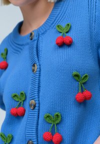 Cardigan en tricot bleu avec cinq applications de cerises en rouge et vert, et des boutons en bois sur le devant. Col texturé à côtes.