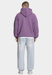 Fioletowa oversizeowa bluza z kapturem na sznurki, zestawiona z jasnoniebieskimi luźnymi dżinsami i białymi trampkami, widoczna od tyłu.