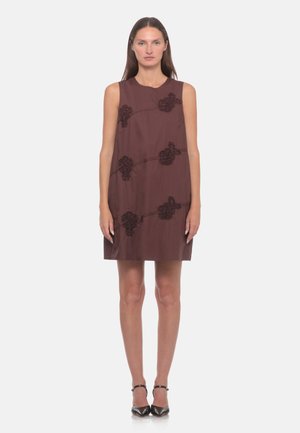 Donna in piedi che indossa un vestito marrone senza maniche con ricamo floreale nero, e tacchi neri con punta a punta e cinturino alla caviglia su sfondo bianco.