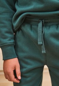 Sweat-shirt en coton bleu sarcelle et pantalon de jogging assorti avec taille élastique, détails à cordon, et poignets côtelés. Texture douce, couture minimale.