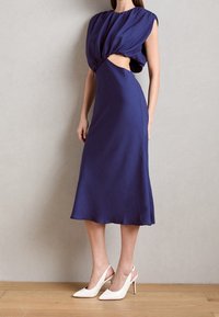 Mörkblå midi-klänning med draperad bodice, utskurna detaljer och slät textur. Kombinerad med vita slingback-höga klackar.