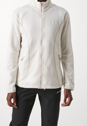 Persoon met een witte fleecejas met rits en twee zakken en een zwarte broek, staand tegen een effen lichte achtergrond.
