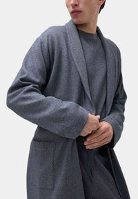 Accappatoio grigio texturizzato con collarino, maniche lunghe e tasche laterali. Indossato sopra una maglietta grigia coordinata, caratterizzata da un design liscio e aderente.