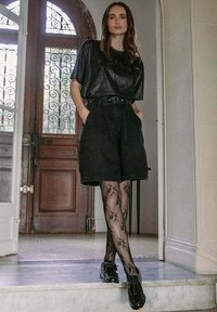 Haut en cuir noir, short en jean noir taille haute, collants noirs à motifs, et chaussures noires brillantes ; debout dans un espace intérieur bien éclairé.