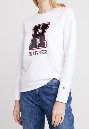 Hvid langærmet sweatshirt med stort sort og rødt bogstav H og "HILFIGER" tekst, båret med blå jeans på en stående person.