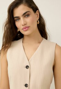 Gilet sans manches beige avec des rayures verticales, présentant un décolleté en V profond et deux boutons noirs à l'avant. Texture lisse, coupe ajustée.