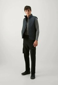 Čierny prešívaný vest s vysokým golierom, nosený cez priliehavý šedý dlhý rukáv. Kombinovaný s čiernymi cargo nohavicami a tmavými topánkami. Jednoduchý dizajn.