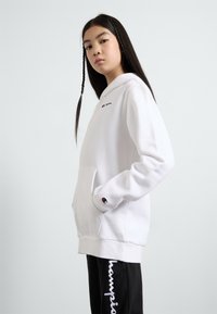 Felpa bianca con cappuccio e tasca frontale, un piccolo logo sul petto e dettagli neri sui polsini. Indossata con pantaloni della tuta neri con testo bianco.