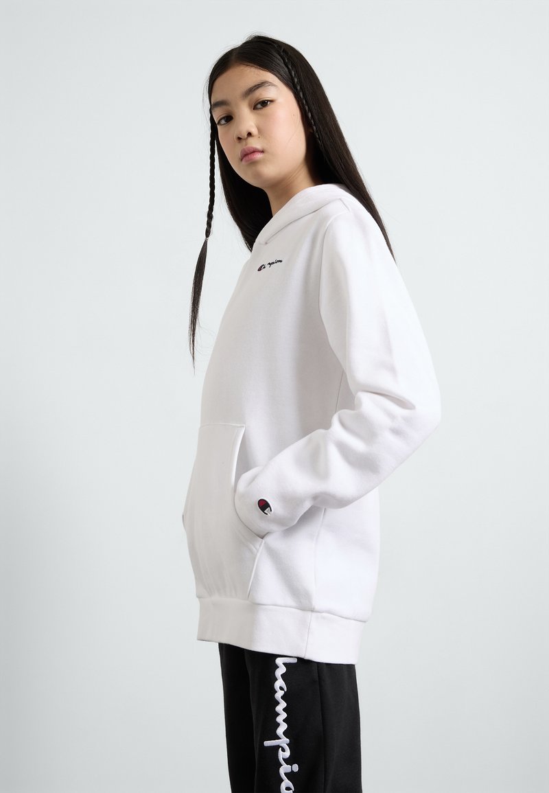 Felpa bianca con cappuccio e tasca frontale, un piccolo logo sul petto e dettagli neri sui polsini. Indossata con pantaloni della tuta neri con testo bianco.