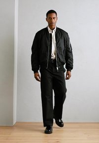 Schwarze Bomberjacke mit Reißverschluss, getragen über einem weißen Hemd und schwarzen Hosen, kombiniert mit schwarzen Schuhen. Das Modell steht vor einer hellen Wand.