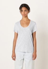 Jauna moteris su garbanotais plaukais, vilkinti šviesiai pilką trumpomis rankovėmis marškinėlį su iškirpte ir kreminės spalvos džinsus, stovinti priešais paprastą foną.