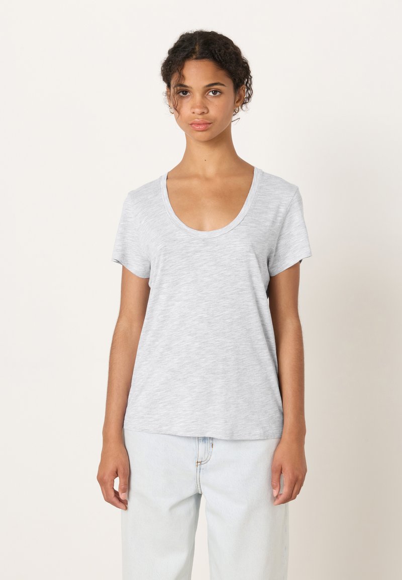 Jauna moteris su garbanotais plaukais, vilkinti šviesiai pilką trumpomis rankovėmis marškinėlį su iškirpte ir kreminės spalvos džinsus, stovinti priešais paprastą foną.