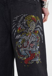 Schwarze Denim-Jeans mit einem bunten Strass-Graphic auf der Gesäßtasche, das komplizierte Muster in Rot, Gelb, Blau und Weiß zeigt.