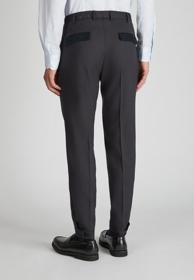 Donkergrijze broeken met een getailleerde pasvorm, voorzien van twee achterzakken met contrasterende textuurpanelen, gedragen met zwarte schoenen.