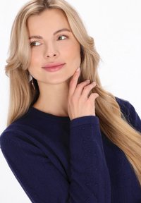 Pull en tricot bleu marine avec un motif texturé, encolure ronde et manches longues. Le modèle touche son menton, affichant un maquillage neutre.