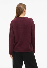 Frau mit langen braunen Haaren, die einen lockeren, dunklen lila Pullover und schwarze Hosen trägt, mit dem Rücken zu einer einfarbigen weißen Hintergrund.