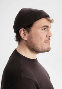 Bruine gebreide muts met een geribbelde textuur, gedragen op het hoofd. De persoon is gekleed in een donkere trui met een eenvoudige ronde hals.