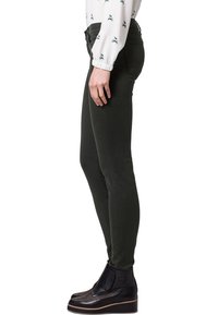 Jeans skinny de color verde oscuro, confeccionados en tela elástica, con cintura alta y bolsillos laterales. Combinados con una blusa blanca con estampado y botas de tobillo negras.