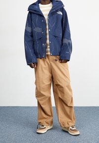 Námořnická voděodolná bunda s motivem blesků, zapínáním na zip a kapucí. Kombinována s bé žovými volnými kalhotami a béžovo-hnědými teniskami.