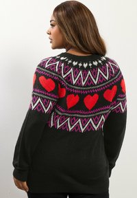 Pull noir avec un motif Fair Isle comprenant des coeurs rouges, des zigzags blancs et roses, et une finition côtelée. Manches longues et col rond.