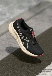 Chaussure de sport noire avec une tige synthétique texturée, accents blanc et rose clair, bout rond et semelle beige rembourrée.