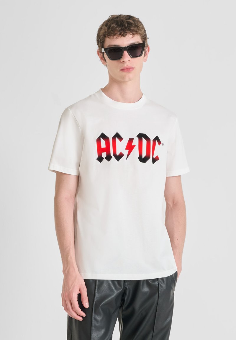 Camiseta blanca de algodón con un audaz logotipo gráfico en rojo y negro de AC/DC. Combinada con pantalones de cuero sintético negros y gafas de sol oscuras.