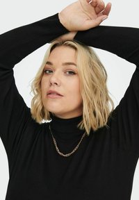Črn, rebrast turtleneck pulover z dolgimi rokavi. Ima zlat verižni nakit. Ozadje je enostavno belo, kar poudarja oblačilo in nakit.
