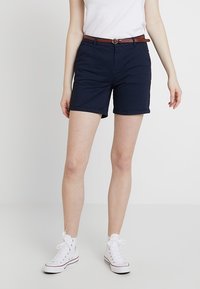 Mörkblå shorts med slim fit, upprullad fåll och ett brunt bälte. Kombineras med vita hög-top sneakers och en enkel vit t-shirt.