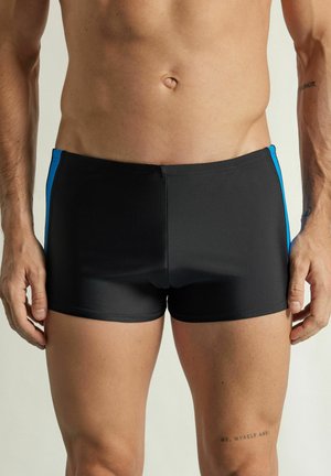 Męski tors i górne partie nóg w czarnych kąpielówkach z niebieskimi bocznymi paskami, stojący na tle jednolitego jasnego tła.