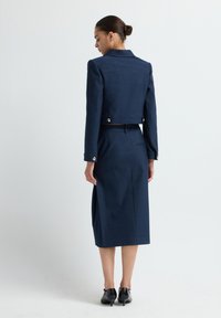 Žena stojí, oblečená v námořnicky modré zkrácené bundě a odpovídající midi sukni s černými nízkými podpatky, na jednoduchém bílém pozadí.