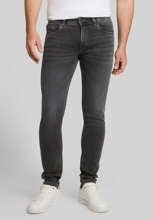 Homme portant un jean gris foncé ajusté et des baskets blanches, debout devant un fond uni.