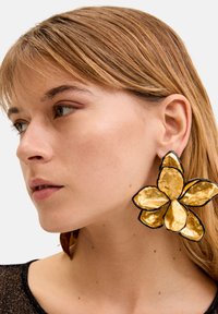 Boucles d'oreilles florales dorées avec des pétales texturés et des bords noirs, présentant un grand design qui pend sous le lobule de l'oreille. Surface reflétant la lumière.