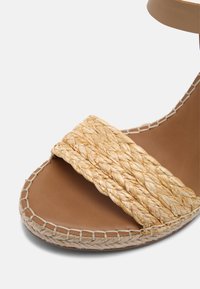 Sandale marron à bout ouvert avec une large bride en paille tressée et une couture tissée le long du bord de la semelle.