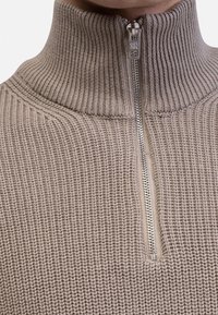 Maglione lavorato a maglia beige con colletto alto e mezza cerniera. Tessuto strutturato con righe verticali. Cerniera argentata con un pratico tirante minimalista.