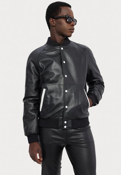 Black leather bomber jacket with white snap buttons and side pockets. Features ribbed cuffs and hem. Smooth texture and streamlined design.