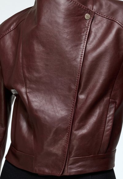 Burgundy leather jacket with a high collar, asymmetrical front, and metal snap closure. Smooth texture and tailored fit with side pockets.
