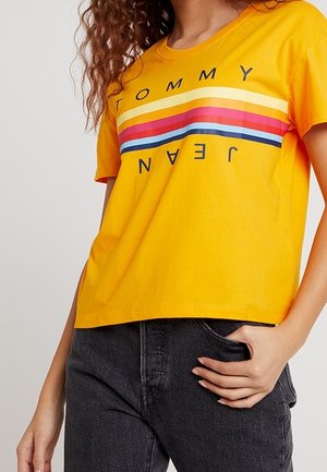 Żółta bawełniana koszulka z poziomym paskowanym wzorem w kolorach czerwonym, niebieskim i białym, z czarnym nadrukiem tekstu "TOMMY" i "JEAN".