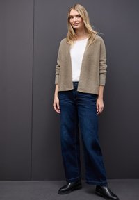 Béžový huňatý cardigan přes bílý top, doplněný tmavě modrými wide-leg džíny a černými kotníkovými botami. Jednoduchý design, neutrální barevná paleta.