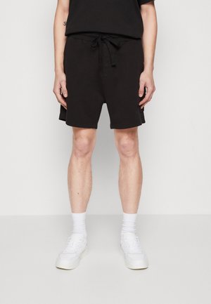 Uomo con pantaloncini neri con coulisse, calzini bianchi e scarpe da ginnastica bianche, in piedi davanti a uno sfondo chiaro e uniforme.