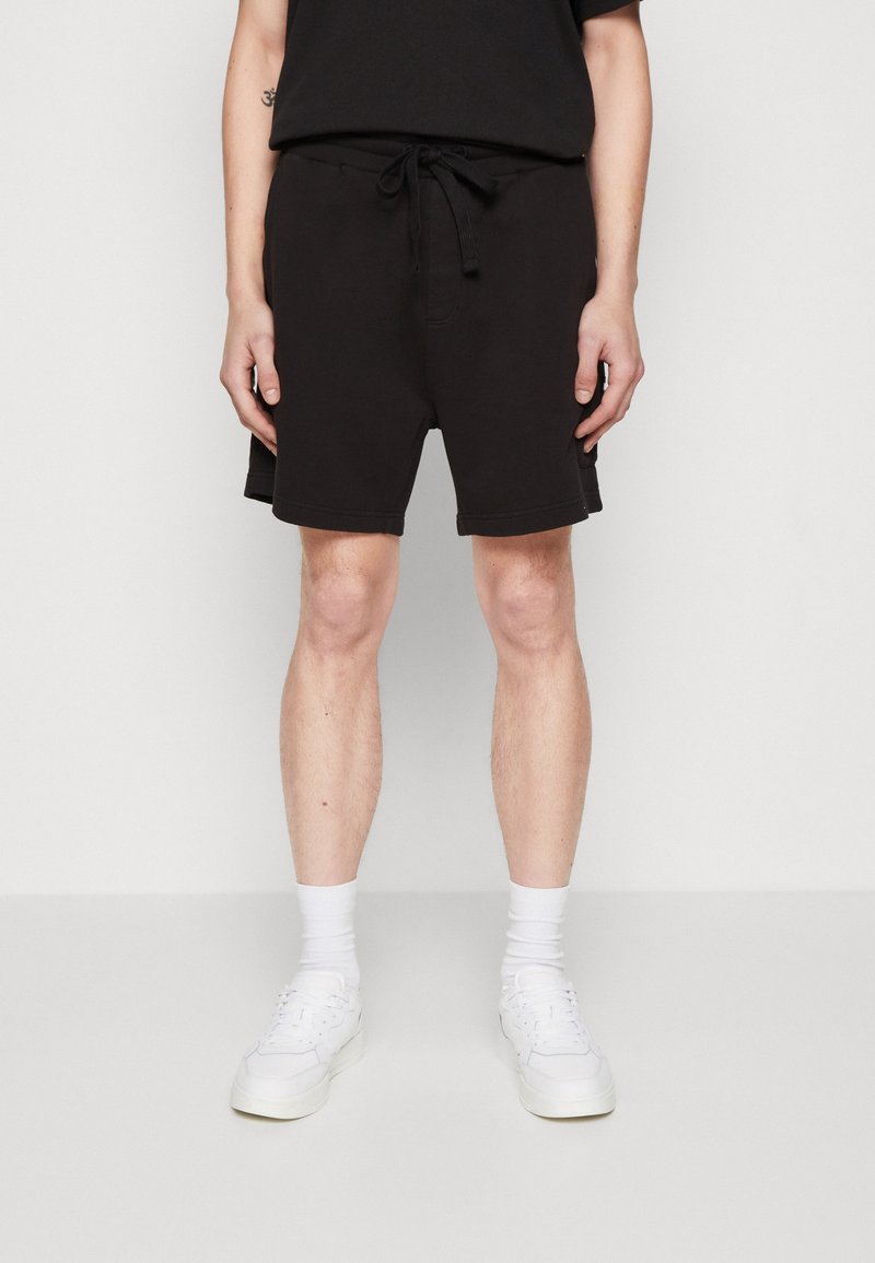 Homme portant un short noir à cordon, des chaussettes blanches montantes et des baskets blanches, debout devant un fond clair uni.
