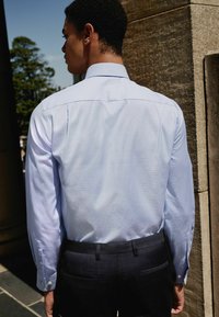 Helle blaue Hemdjacke mit langen Ärmeln, sichtbarem Kragen und Manschetten, kombiniert mit dunklen Hosen mit Taschen. Strukturierte Oberfläche.
