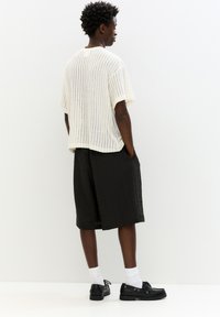 Maglietta bianca a righe con maniche corte, abbinata a pantaloni corti neri con texture. Modello che indossa scarpe nere e calze bianche, visto da dietro.