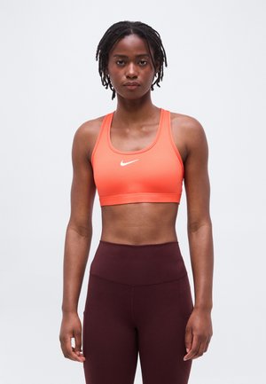 Femeie atletica cu dreadlocks scurte, purtând un sutien sport portocaliu Nike și colanți lungi înalți de culoare maro închis, stând în picioare pe un fundal simplu.