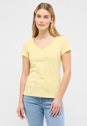 Jeune femme aux longs cheveux blonds portant un t-shirt jaune clair à manches courtes et col en V, ainsi qu'un jean bleu clair, debout devant un fond uni.