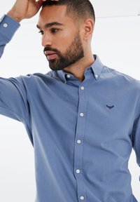 Jasnoniebieska koszula zapinana na guziki, wykonana z bawełny. Posiada kołnierz z ostrymi końcami, długie rękawy oraz mały haftowany emblemat na piersi.