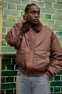 Brown bomber jacket with a shearling collar, featuring two front pockets and ribbed cuffs. Worn with light blue denim jeans. Green tiled background.