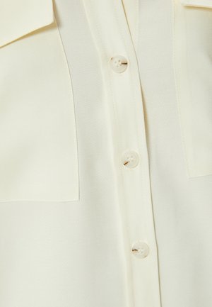Camicia beige chiaro con tessuto texturizzato, due tasche sul petto e bottoni sulla parte anteriore. Presenta un design colletto classico.